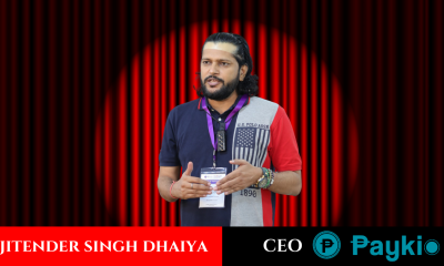 Jitender Dhaiya, CEO Paykio dressed in a casual t-shirt and blazer, speaks at a podium against a red striped background.