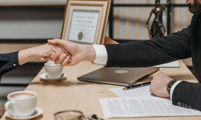 Two individuals engaging in a firm handshake over a desk with paperwork and coffee cups visible.